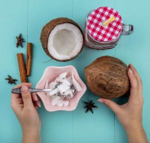 top-view-female-hands-holding-one-hand-spoon-with-pulps-coconut-other-hand-whole-coconut-with-cinnamonnise-glass-jar-blue